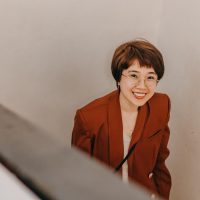 编辑林小满的插画头像，短发戴圆框眼镜，手持铅笔在关卡草图旁做批注，背景是奶油色与砖红色块